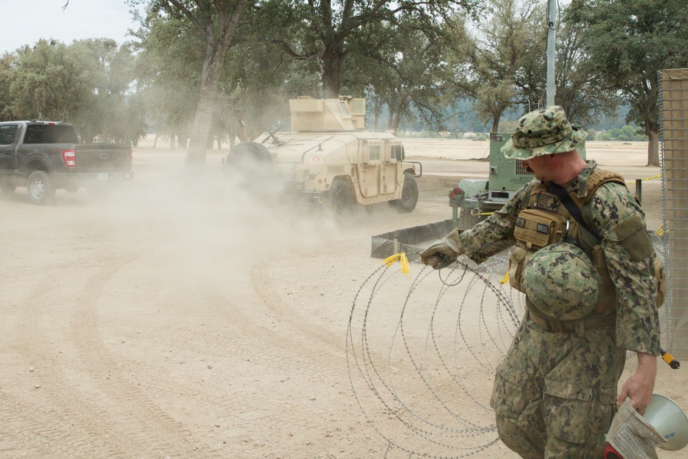 A U.S. Navy sailor in NWU Type III (woodland digital camouflage) uniform is handling concertina/razor wire at what appears to be a military training or operational base. In the background, a tan-colored HMMWV (Humvee) and a civilian pickup truck are visible amid a dust cloud on a dirt road. The setting appears to be an arid, semi-wooded training environment.