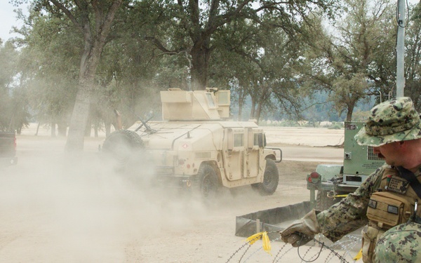 A U.S. Navy sailor in NWU Type III (woodland digital camouflage) uniform is handling concertina/razor wire at what appears to be a military training or operational base. In the background, a tan-colored HMMWV (Humvee) and a civilian pickup truck are visible amid a dust cloud on a dirt road. The setting appears to be an arid, semi-wooded training environment.