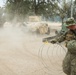 A U.S. Navy sailor in NWU Type III (woodland digital camouflage) uniform is handling concertina/razor wire at what appears to be a military training or operational base. In the background, a tan-colored HMMWV (Humvee) and a civilian pickup truck are visible amid a dust cloud on a dirt road. The setting appears to be an arid, semi-wooded training environment.