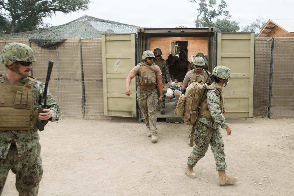 Military personnel in camouflage uniforms are conducting what appears to be a medical evacuation or casualty extraction drill at a forward operating base or training facility. Several service members are carrying a patient on a litter/stretcher out of a modified shipping container that appears to serve as a medical treatment facility. The setting includes sand-covered ground, camouflage netting, and temporary military structures consistent with a field training environment.