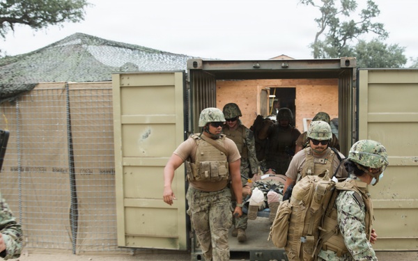Military personnel in camouflage uniforms are conducting what appears to be a medical evacuation or casualty extraction drill at a forward operating base or training facility. Several service members are carrying a patient on a litter/stretcher out of a modified shipping container that appears to serve as a medical treatment facility. The setting includes sand-covered ground, camouflage netting, and temporary military structures consistent with a field training environment.