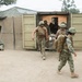 Military personnel in camouflage uniforms are conducting what appears to be a medical evacuation or casualty extraction drill at a forward operating base or training facility. Several service members are carrying a patient on a litter/stretcher out of a modified shipping container that appears to serve as a medical treatment facility. The setting includes sand-covered ground, camouflage netting, and temporary military structures consistent with a field training environment.