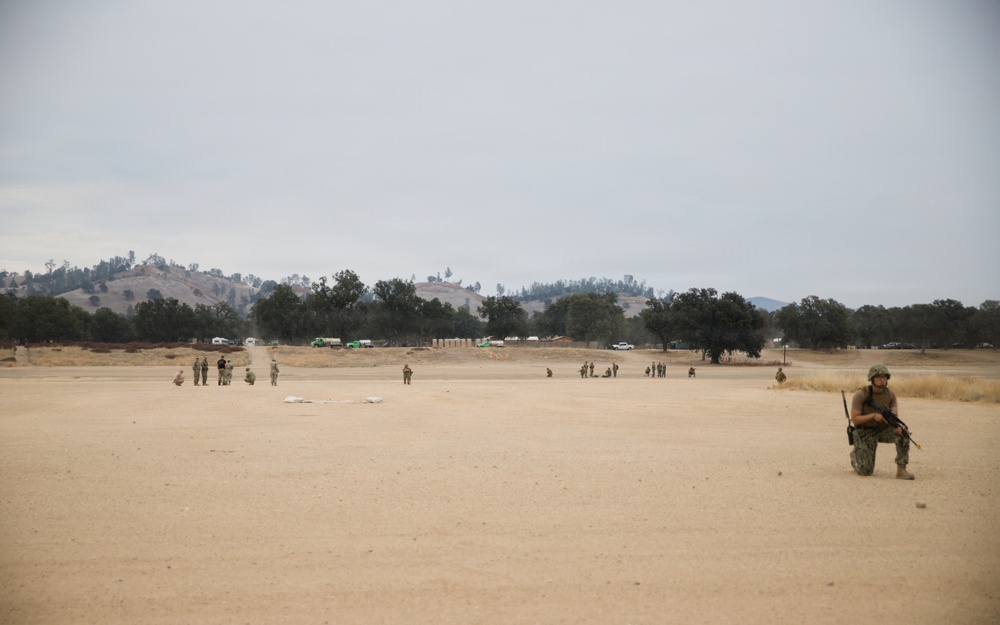 The image shows a military training exercise taking place on a large open sandy field. Multiple personnel in camouflage uniforms are dispersed across the area, with one soldier prominently kneeling in the foreground holding a rifle. The background features rolling hills, oak trees, and several military vehicles, consistent with a training base environment.