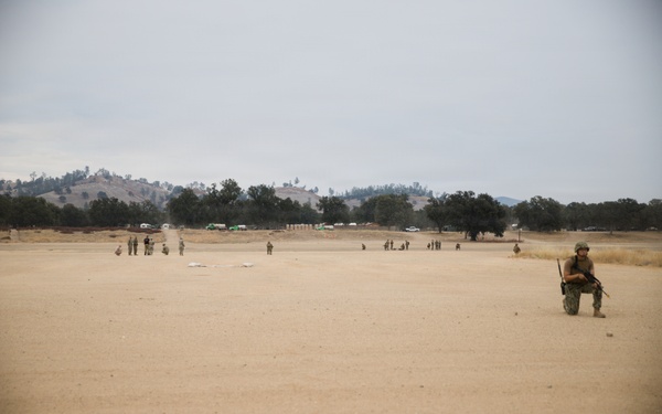 The image shows a military training exercise taking place on a large open sandy field. Multiple personnel in camouflage uniforms are dispersed across the area, with one soldier prominently kneeling in the foreground holding a rifle. The background features rolling hills, oak trees, and several military vehicles, consistent with a training base environment.