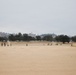 The image shows a military training exercise taking place on a large open sandy field. Multiple personnel in camouflage uniforms are dispersed across the area, with one soldier prominently kneeling in the foreground holding a rifle. The background features rolling hills, oak trees, and several military vehicles, consistent with a training base environment.