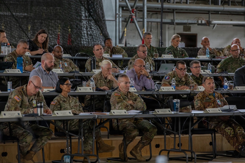 A large military briefing or conference is taking place inside what appears to be a hangar or large industrial facility. Multiple rows of military personnel in various camouflage uniforms, along with a few civilians, are seated at long tables facing a presenter or screen outside the frame. Name placards are visible on the tables, indicating a formal, organized meeting.