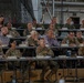 A large military briefing or conference is taking place inside what appears to be a hangar or large industrial facility. Multiple rows of military personnel in various camouflage uniforms, along with a few civilians, are seated at long tables facing a presenter or screen outside the frame. Name placards are visible on the tables, indicating a formal, organized meeting.