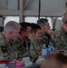 A group of military personnel from multiple nations are seated at a table during what appears to be a briefing or conference in a field/hangar setting. At least two soldiers on the left wear Lithuanian flag patches on their camouflage uniforms, while others display US Army patches. The group appears attentive and engaged, suggesting a multinational military coordination meeting.