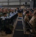 A large military conference or briefing is taking place inside what appears to be a hangar or large indoor facility. Numerous military personnel from multiple nations, wearing various camouflage uniforms, are seated at long tables arranged in rows facing a common direction. The setting suggests a multinational military exercise, summit, or joint operations planning event.