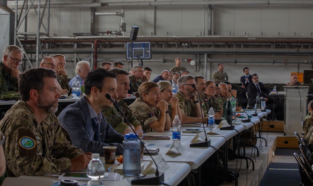 A formal military briefing or conference is taking place inside what appears to be a large hangar or warehouse facility. Multiple military personnel in various uniforms and a few civilians in business attire are seated at long tables facing a presenter or screen off-camera. The setting suggests a multinational military meeting or exercise coordination event.