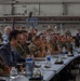 A formal military briefing or conference is taking place inside what appears to be a large hangar or warehouse facility. Multiple military personnel in various uniforms and a few civilians in business attire are seated at long tables facing a presenter or screen off-camera. The setting suggests a multinational military meeting or exercise coordination event.