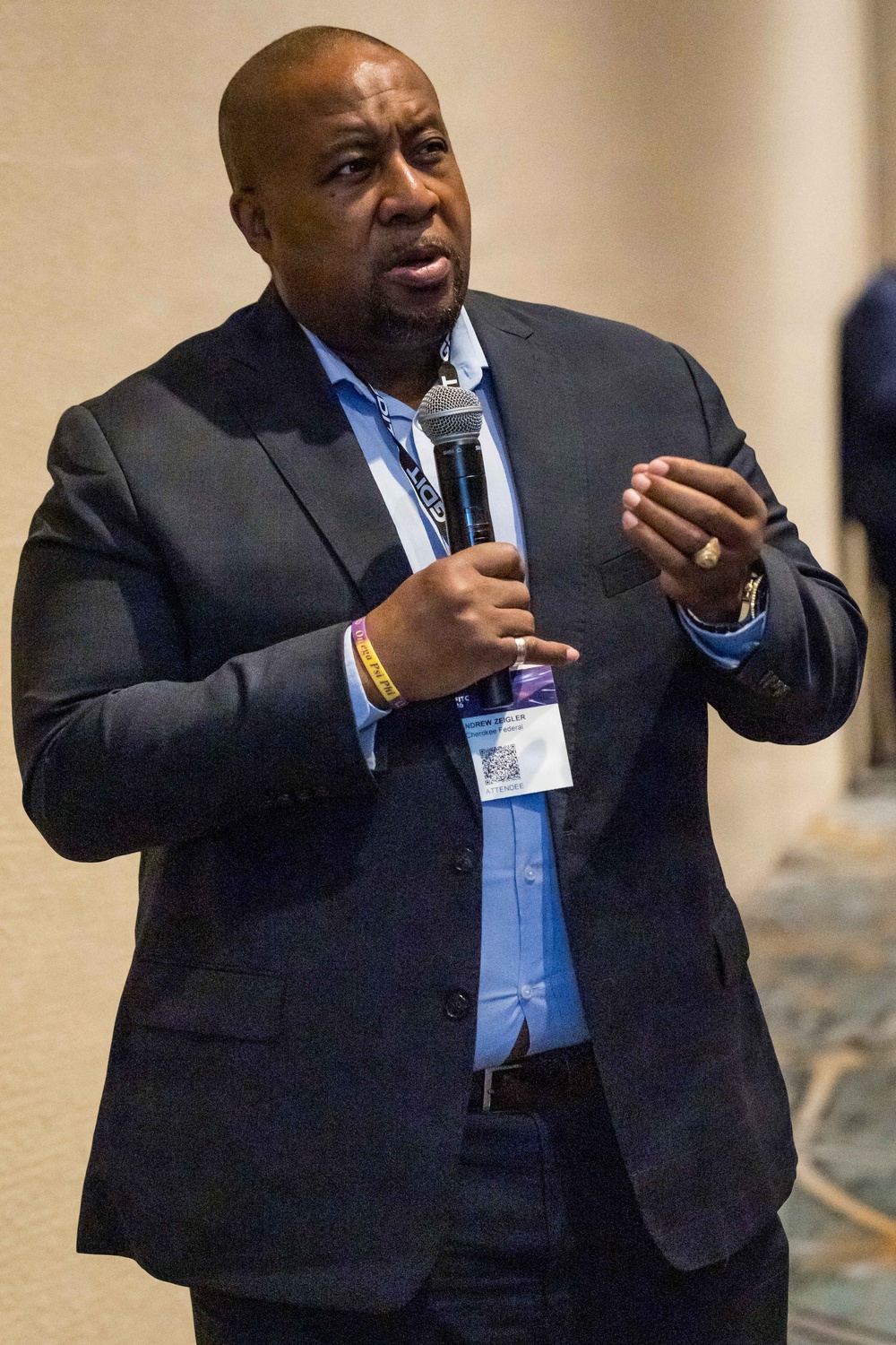 A man is speaking into a handheld microphone at what appears to be a professional conference or event. He is wearing a dark suit with a light blue dress shirt and a conference badge on a lanyard. His right hand is raised in a gesturing motion, suggesting he is actively presenting or addressing an audience.