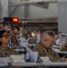 A military briefing or conference is taking place in what appears to be a large hangar or industrial facility. Multiple U.S. Army personnel in OCP (Operational Camouflage Pattern) uniforms are seated at tables with documents, microphones, and water bottles. The foreground features two soldiers prominently, one with visible 'Old Ironsides' (1st Armored Division) unit patch.