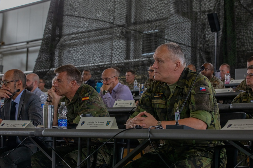 A multinational military briefing or conference is taking place in what appears to be a military hangar or field facility decorated with camouflage netting. Several senior military officers from different nations, including Spain and the Czech Republic, are seated at tables listening attentively. Name placards are visible on the tables, identifying at least one attendee as Maj Gen Petr Svoboda, Territorial Forces Command Commander.