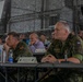 A multinational military briefing or conference is taking place in what appears to be a military hangar or field facility decorated with camouflage netting. Several senior military officers from different nations, including Spain and the Czech Republic, are seated at tables listening attentively. Name placards are visible on the tables, identifying at least one attendee as Maj Gen Petr Svoboda, Territorial Forces Command Commander.