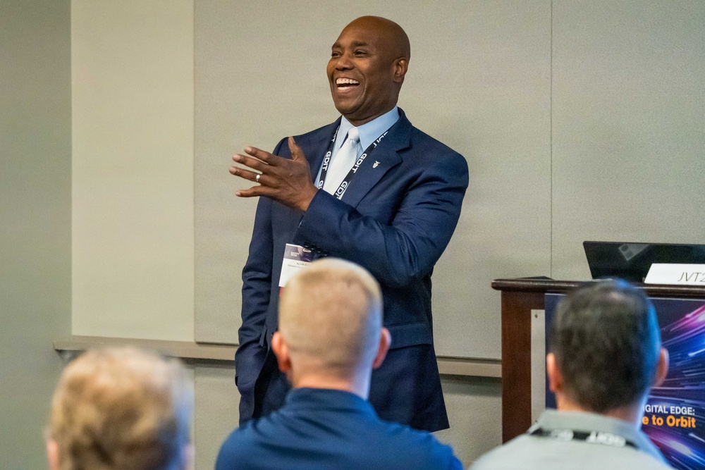 A man in a navy blue suit is presenting or speaking at what appears to be a professional conference or briefing. He is laughing and gesturing with his hands, engaging an audience seated in front of him. A presentation screen partially visible in the lower right shows text including 'DIGITAL EDGE' and 'to Orbit'.