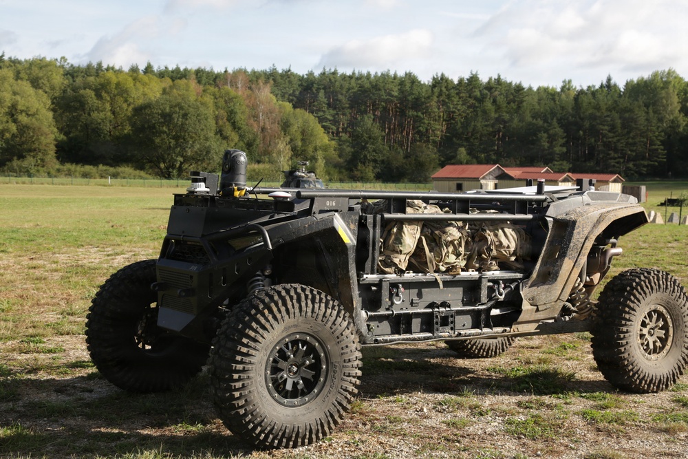 The image shows a military unmanned ground vehicle (UGV) or tactical off-road vehicle parked on a grassy field. The vehicle has an open frame design with large all-terrain tires, sensor/camera mounts on top, and what appears to be military gear stored in the cargo area. A forested background with a building and what may be a helicopter are visible in the distance.