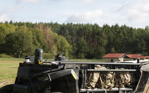 The image shows a military unmanned ground vehicle (UGV) or tactical off-road vehicle parked on a grassy field. The vehicle has an open frame design with large all-terrain tires, sensor/camera mounts on top, and what appears to be military gear stored in the cargo area. A forested background with a building and what may be a helicopter are visible in the distance.