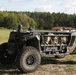 The image shows a military unmanned ground vehicle (UGV) or tactical off-road vehicle parked on a grassy field. The vehicle has an open frame design with large all-terrain tires, sensor/camera mounts on top, and what appears to be military gear stored in the cargo area. A forested background with a building and what may be a helicopter are visible in the distance.