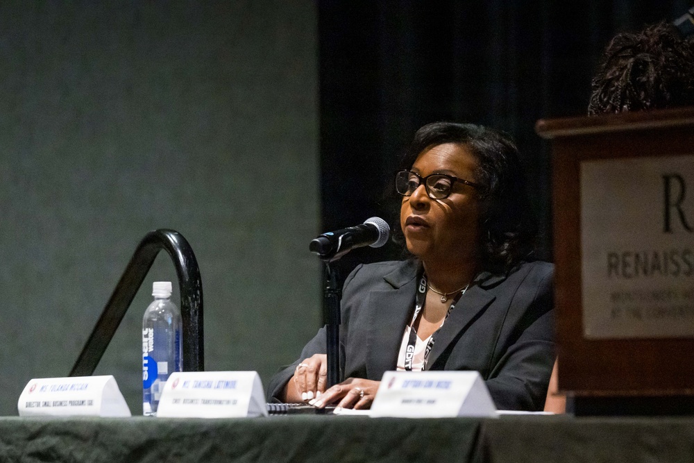 A woman wearing glasses and a dark blazer is speaking at a panel discussion at what appears to be a Renaissance hotel or convention center. She is seated at a table with name placards visible in front of multiple panelists, speaking into a microphone. The setting suggests a professional conference or business event.