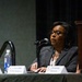 A woman wearing glasses and a dark blazer is speaking at a panel discussion at what appears to be a Renaissance hotel or convention center. She is seated at a table with name placards visible in front of multiple panelists, speaking into a microphone. The setting suggests a professional conference or business event.