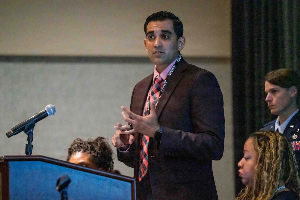 A man in a dark suit with a pink shirt and plaid tie is speaking at a formal event, standing near a podium with a microphone. He is wearing a lanyard that reads 'GDIT' and appears to be addressing an audience. In the background, a woman in military uniform and another individual are visible.