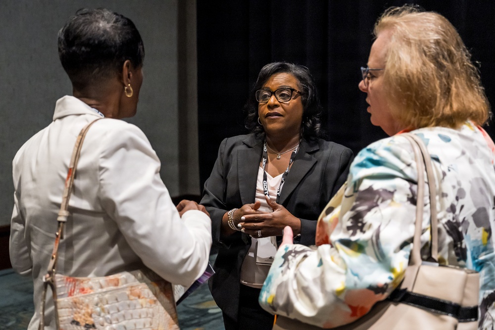 Three women are engaged in conversation at what appears to be a professional conference or event. The central figure, wearing a black blazer and conference lanyard, appears to be speaking to the other two attendees. The setting suggests a networking or post-session discussion in a conference venue.