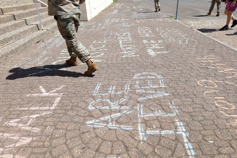 A paved walkway covered in chalk writings and messages, with several people in military camouflage uniforms walking across it. The chalk messages appear to include phrases related to social justice or awareness campaigns. The scene appears to be outdoors near a building with steps visible on the left side.