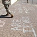 A paved walkway covered in chalk writings and messages, with several people in military camouflage uniforms walking across it. The chalk messages appear to include phrases related to social justice or awareness campaigns. The scene appears to be outdoors near a building with steps visible on the left side.