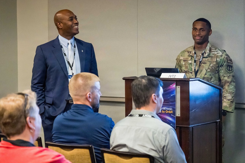 A U.S. Air Force service member in OCP (Operational Camouflage Pattern) uniform stands at a wooden podium labeled 'JVT2' during what appears to be a professional conference or briefing. A civilian in a dark blue suit stands beside him, both smiling toward an audience. The podium displays a graphic reading 'Digital Edge: [Edge] to Orbit,' suggesting a technology-focused military event.