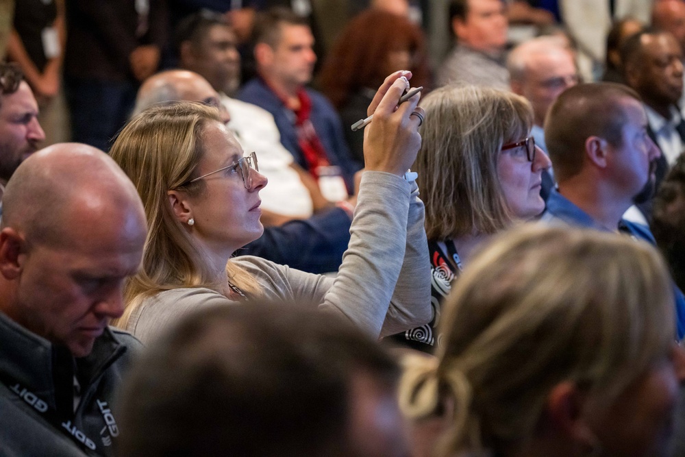A large audience is seated at what appears to be a professional conference or seminar. A woman in the foreground with glasses and blonde hair is raising her hand while holding a pen, likely asking a question or participating in a discussion. The crowd is densely packed and attentive, suggesting an engaged audience at a formal event.