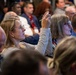 A large audience is seated at what appears to be a professional conference or seminar. A woman in the foreground with glasses and blonde hair is raising her hand while holding a pen, likely asking a question or participating in a discussion. The crowd is densely packed and attentive, suggesting an engaged audience at a formal event.