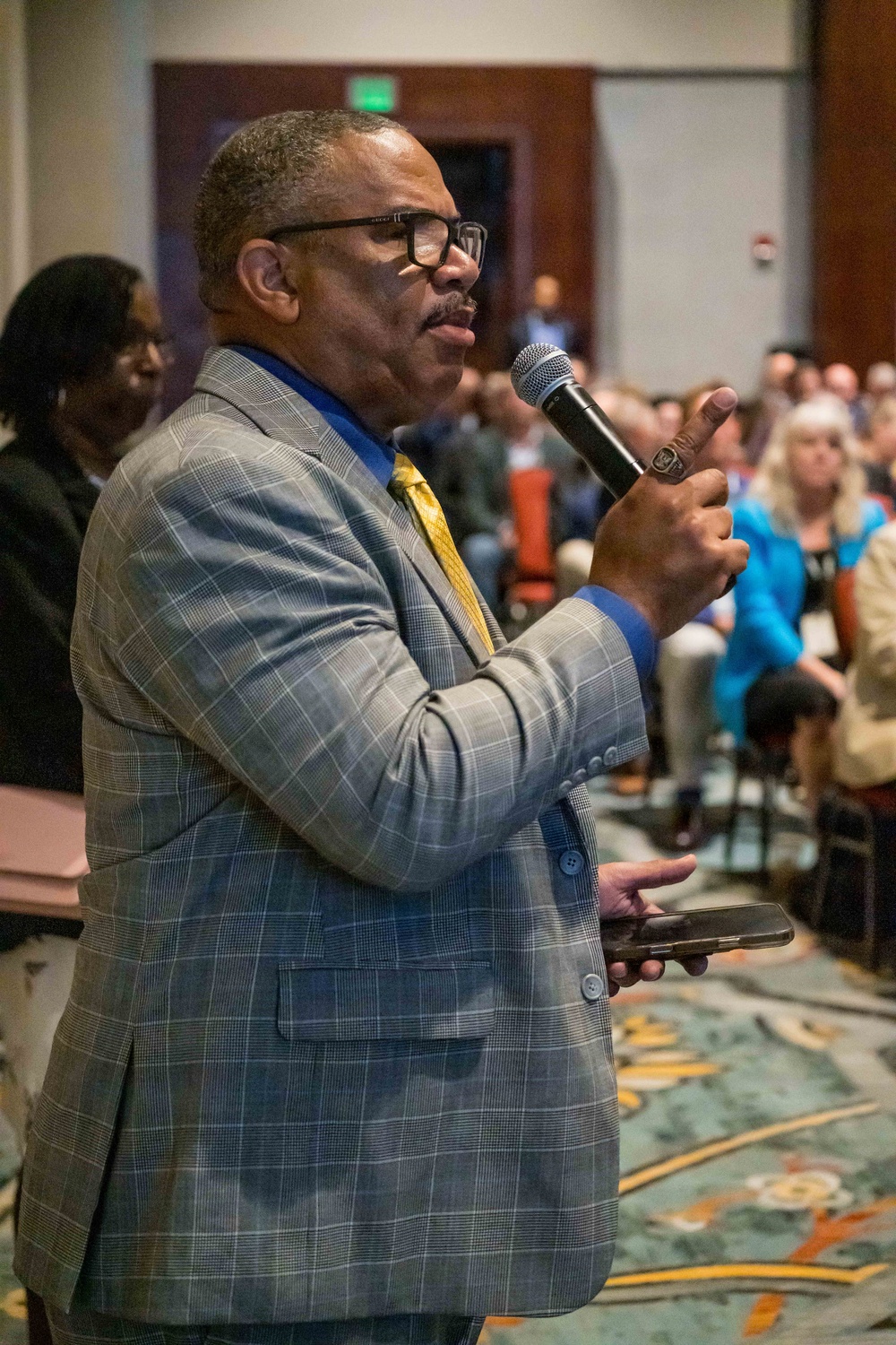A man in a gray plaid suit with a blue dress shirt and gold tie is speaking into a handheld microphone at what appears to be a professional conference or event. He is holding a smartphone in his other hand and gesturing with his microphone hand while addressing a seated audience. The setting is a large conference hall or ballroom with patterned carpeting.
