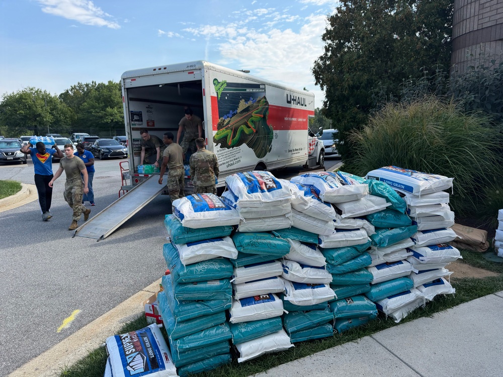 Military personnel and civilians are unloading supplies from a U-Haul moving truck in a parking lot. A large pallet of bagged goods, appearing to be rice or similar bulk food items, is stacked on the grass nearby. This appears to be a disaster relief or humanitarian aid distribution operation.