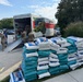 Military personnel and civilians are unloading supplies from a U-Haul moving truck in a parking lot. A large pallet of bagged goods, appearing to be rice or similar bulk food items, is stacked on the grass nearby. This appears to be a disaster relief or humanitarian aid distribution operation.