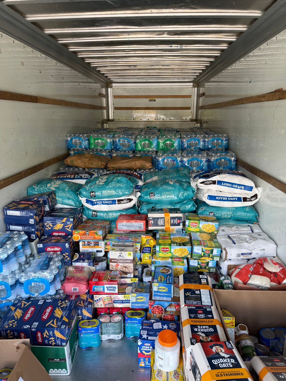 The interior of a large freight trailer is packed floor-to-ceiling with donated or relief food and water supplies. Items include bottled water, rice, pasta, canned goods, oatmeal, and other non-perishable staples, suggesting a disaster relief or food bank distribution effort. The trailer appears nearly full, indicating a large-scale humanitarian supply operation.