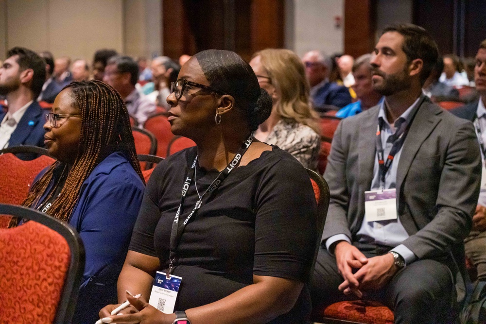 A professional conference or seminar setting with a large audience seated in red upholstered chairs. Three individuals are prominently visible in the foreground, all wearing lanyards with 'GDIT' branding, attentively watching a presentation. The audience appears diverse and professionally dressed, filling a large conference hall.