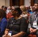 A professional conference or seminar setting with a large audience seated in red upholstered chairs. Three individuals are prominently visible in the foreground, all wearing lanyards with 'GDIT' branding, attentively watching a presentation. The audience appears diverse and professionally dressed, filling a large conference hall.