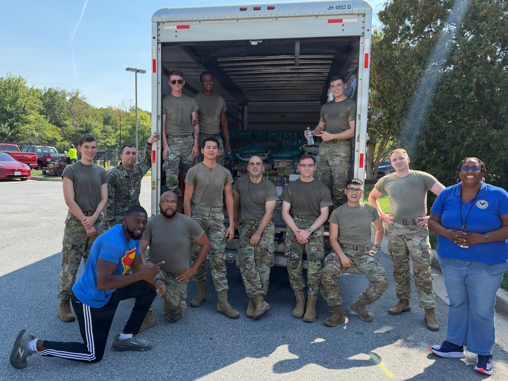 A group of approximately 13 individuals, mostly in military camouflage uniforms (OCP pattern), pose in front of an open box truck loaded with supplies, likely water and food items. Two civilians are present — one in a blue Superman t-shirt kneeling in front and one in a blue polo shirt on the right. The scene appears to be a community service or humanitarian aid effort, possibly a food/supply drive.