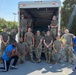A group of approximately 13 individuals, mostly in military camouflage uniforms (OCP pattern), pose in front of an open box truck loaded with supplies, likely water and food items. Two civilians are present — one in a blue Superman t-shirt kneeling in front and one in a blue polo shirt on the right. The scene appears to be a community service or humanitarian aid effort, possibly a food/supply drive.