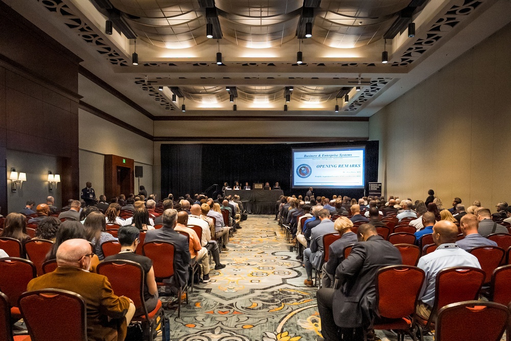 A large conference or symposium is underway in a hotel ballroom, with a substantial audience seated in red chairs facing a panel table at the front. A projection screen displays 'Business &amp; Enterprise Systems - Opening Remarks,' indicating this is the beginning of a formal professional or government-related event. The room is well-attended with approximately 150-200 attendees dressed in business and business-casual attire.