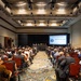 A large conference or symposium is underway in a hotel ballroom, with a substantial audience seated in red chairs facing a panel table at the front. A projection screen displays 'Business &amp; Enterprise Systems - Opening Remarks,' indicating this is the beginning of a formal professional or government-related event. The room is well-attended with approximately 150-200 attendees dressed in business and business-casual attire.