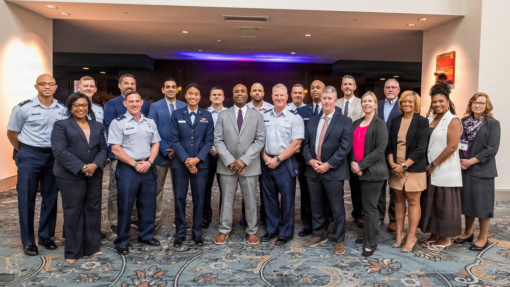 A formal group photograph of approximately 22 individuals posed together in what appears to be a hotel ballroom or conference venue. The group includes a mix of U.S. Air Force military personnel in uniform and civilians in business attire. The composition suggests an official event, ceremony, or professional gathering.