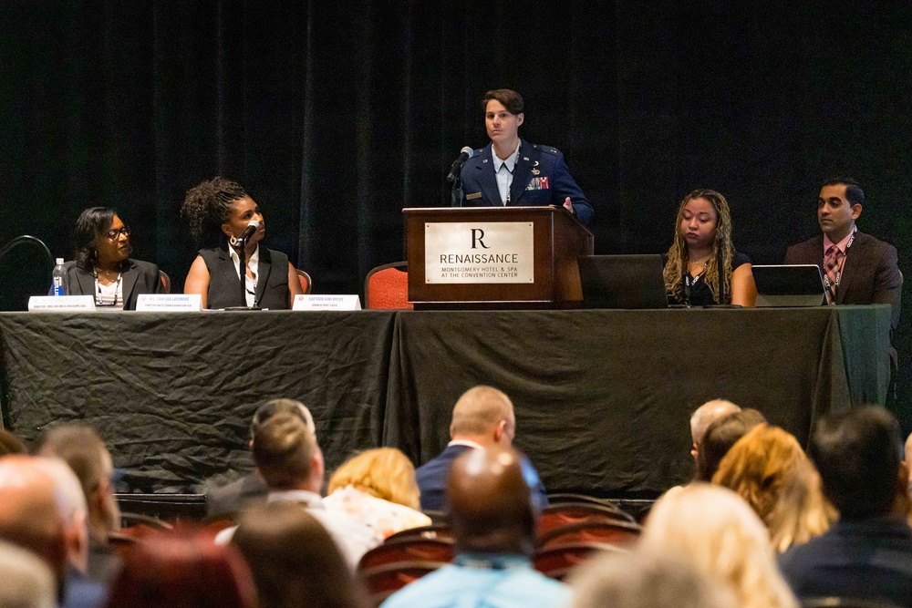A panel discussion is taking place at the Renaissance Montgomery Hotel &amp; Spa at the Convention Center. A military officer in dress uniform stands at the podium while four other panelists are seated at a draped table on either side. An audience is visible in the foreground attending the event.