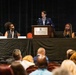 A panel discussion is taking place at the Renaissance Montgomery Hotel &amp; Spa at the Convention Center. A military officer in dress uniform stands at the podium while four other panelists are seated at a draped table on either side. An audience is visible in the foreground attending the event.
