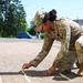 A female U.S. Army soldier in Operational Camouflage Pattern (OCP) uniform is crouched down on a paved sidewalk, using chalk to mark or write something on the ground. The setting appears to be a military installation or base, with a road, vehicles, and trees visible in the background. The activity suggests a training exercise, inspection marking, or administrative task.