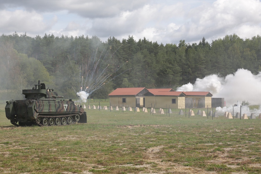 A military training exercise is underway on an open field, featuring an armored personnel carrier firing pyrotechnics or flares toward a simulated village or target area. White smoke and sparks are visible near mock buildings with red roofs, suggesting a live-fire or demonstration drill. The scene includes defensive obstacles and a treeline in the background under a partly cloudy sky.