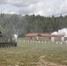 A military training exercise is underway on an open field, featuring an armored personnel carrier firing pyrotechnics or flares toward a simulated village or target area. White smoke and sparks are visible near mock buildings with red roofs, suggesting a live-fire or demonstration drill. The scene includes defensive obstacles and a treeline in the background under a partly cloudy sky.