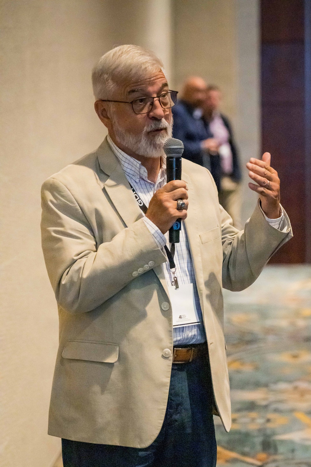 An older man with white/gray hair and a beard is speaking into a handheld microphone at what appears to be a professional conference or event. He is wearing a beige blazer, a plaid shirt, dark jeans, and a conference lanyard with a name badge. His right hand is raised in a gesturing motion, suggesting active engagement with an audience.