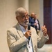 An older man with white/gray hair and a beard is speaking into a handheld microphone at what appears to be a professional conference or event. He is wearing a beige blazer, a plaid shirt, dark jeans, and a conference lanyard with a name badge. His right hand is raised in a gesturing motion, suggesting active engagement with an audience.